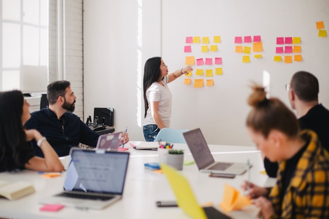 Woman placing sticky notes on a white wall while colleagues watch from a conference table with laptops.